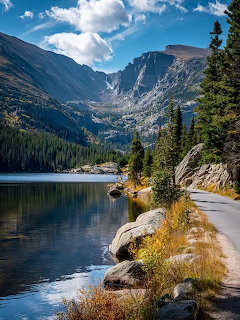 Mountain lake beside a narrow road with pine trees, rocks, and peaks under a blue sky