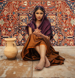 Woman in burgundy and gold robes sitting before an intricate patterned carpet with a clay pot