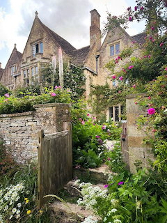 Stone cottage with climbing roses and lush garden flowers behind a rustic wooden gate