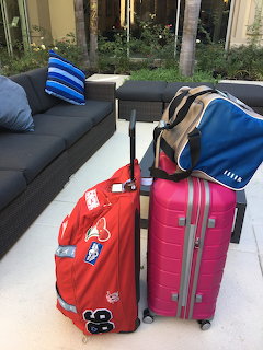 Red and pink luggage with a blue duffel bag beside an outdoor sofa on a patio