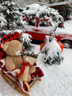 Teddy bear sitting on wooden sled in snow with red truck and Christmas trees behind