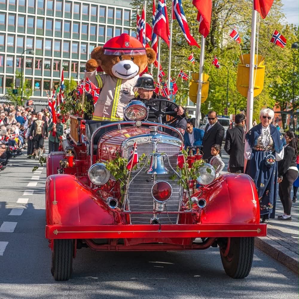 Red vintage fire truck decorated with Norwegian flags and ornaments in a street parade with spectators