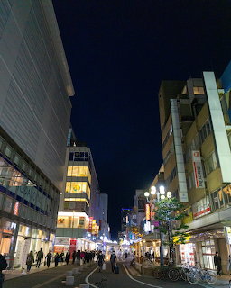 Night city street with lit buildings, streetlights, pedestrians, and bicycles