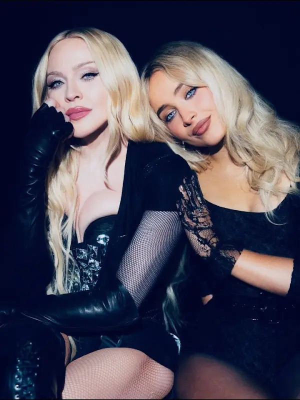 Two blonde women in black outfits posing together against a dark background