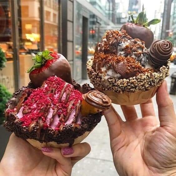 Two hands holding extravagantly decorated donuts with chocolate, candy, strawberries, and various toppings