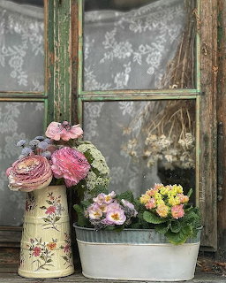Pink flowers in vintage vase and small blooms in white pot against old weathered window with peeling green shutters