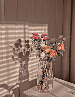 Orange and pink roses in a clear glass vase on a white table with striped sunlight on the wall
