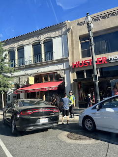 Sunny city street with parked cars, pedestrians, and storefronts in a low-rise building