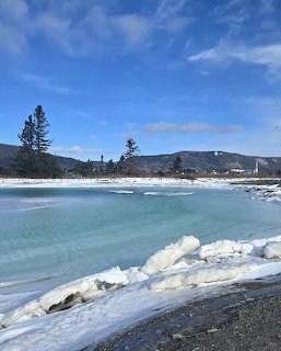 Frozen shoreline with ice chunks, turquoise water, and distant snow-covered mountains under a blue sky