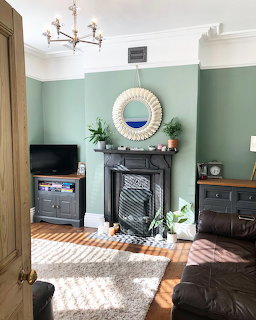 Living room with sage green walls, black fireplace, round mirror, TV stand, plants, and leather sofa