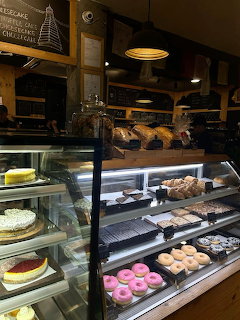 Glass display case in a café showing colorful donuts, cakes, and pastries with a chalkboard menu behind