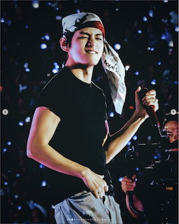 Young man performing on stage in a black shirt and backward cap with blurred concert lights behind him