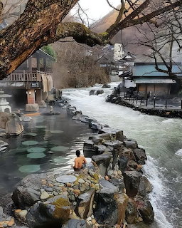 Person bathing in outdoor onsen hot spring next to a flowing river with traditional Japanese buildings and mountains