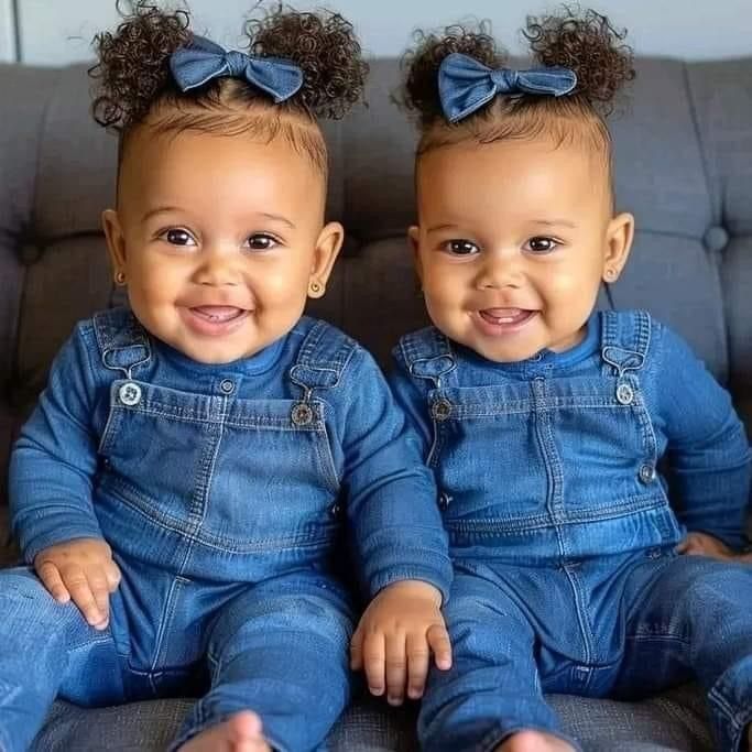 Two smiling baby girls in matching denim clothes and blue bows sitting on a couch