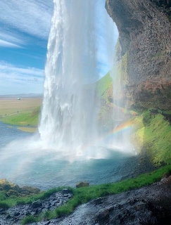 Tall waterfall pouring over rocky cliff with rainbow in mist and green landscape behind
