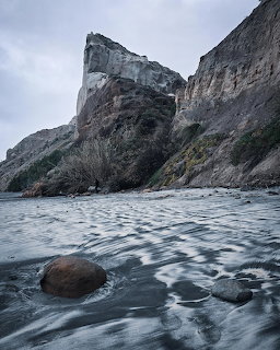 Tall rocky cliffs rise above a sandy beach with waves under gray cloudy skies