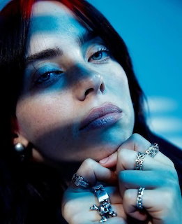 Close-up portrait of Billie Eilish with blue-toned lighting, wearing silver rings and a cross pendant