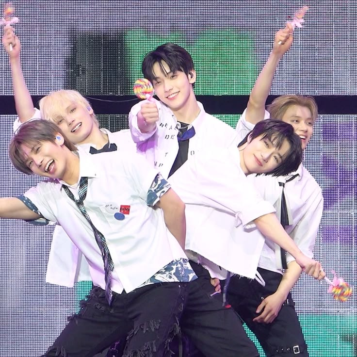 Five K-pop performers in white shirts posing with raised arms on a colorful stage