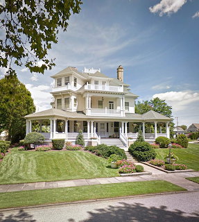 White three-story Victorian house with wrap porch, green lawn, and trees