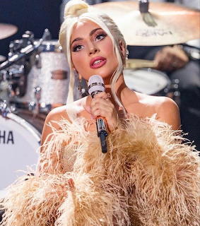 Blonde woman in beige feathered outfit singing into microphone on stage