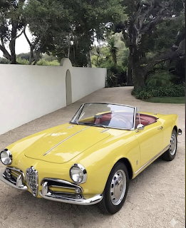 Yellow vintage Alfa Romeo Spider convertible parked on a driveway near a white wall and trees