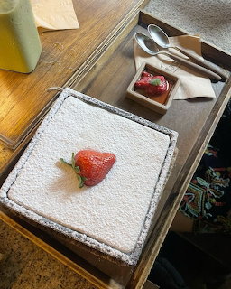 Square powdered cake topped with a strawberry on a wooden table with utensils and a small dessert tray
