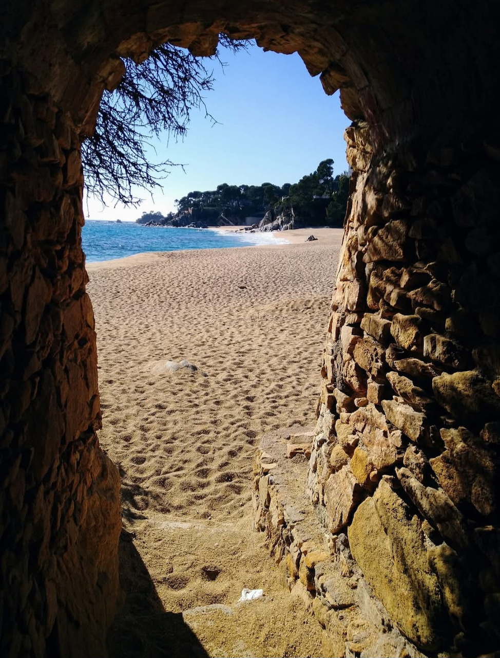 View through a stone arch tunnel opening onto a sandy beach and calm blue sea