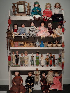 Vintage dolls displayed on four white shelves with decorative mirror frame