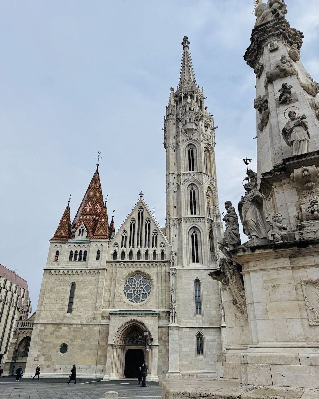 White Gothic church with tall spire and red-roofed tower next to ornate Trinity Column in Budapest