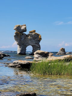 Weathered limestone sea stack with natural arch opening standing in shallow coastal water against blue sky