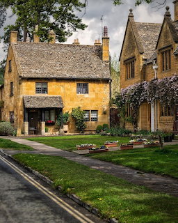 Golden stone cottages beside a quiet village road with green lawns and blooming trees