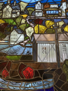 Stained glass window showing a village scene with houses, churches, autumn trees, and a river