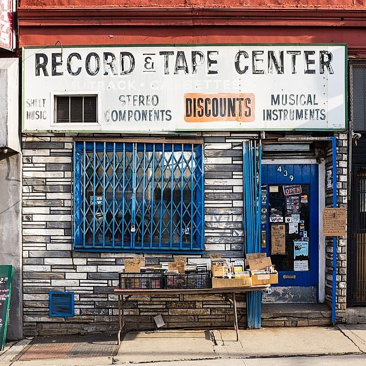 Vintage record and tape center storefront with blue security gate and wooden bench on sidewalk