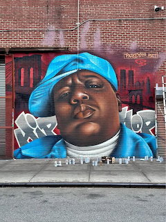 Street mural portrait of Biggie on a red brick wall above a city sidewalk