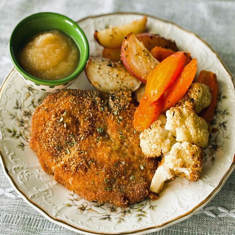 Breaded pork chop dinner with roasted potatoes, carrots, cauliflower, and applesauce on a plate
