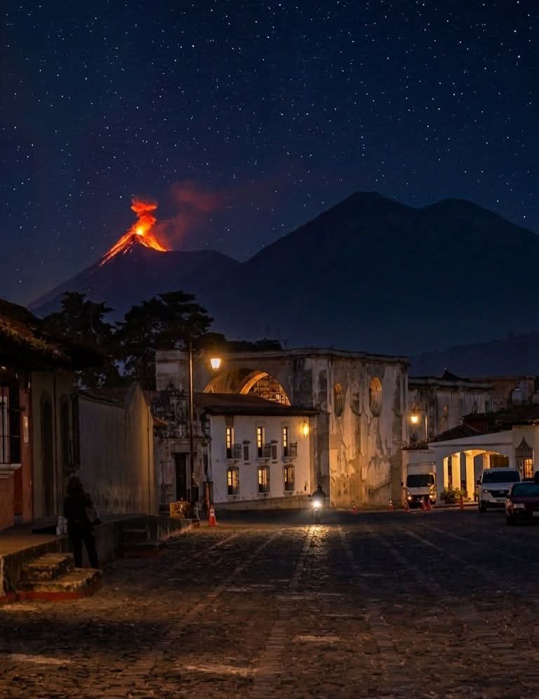 Night street scene in Antigua Guatemala with colonial buildings and an erupting volcano glowing under a starry sky