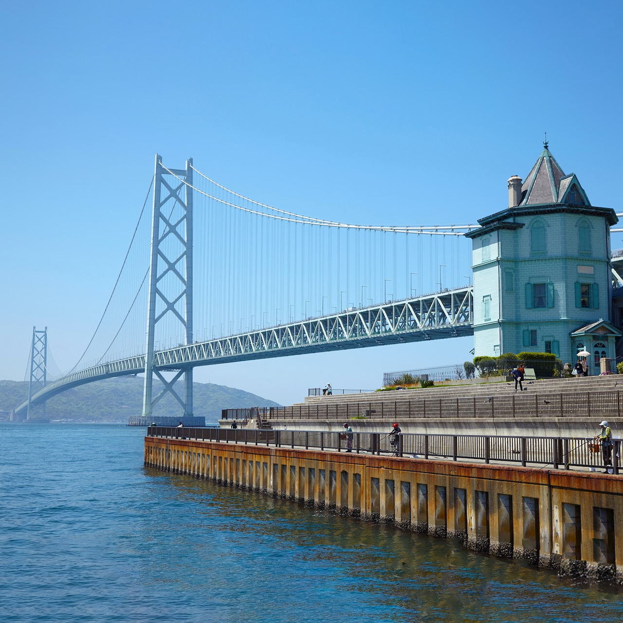Akashi Kaikyo suspension bridge over blue water with turquoise coastal building and wooden pier