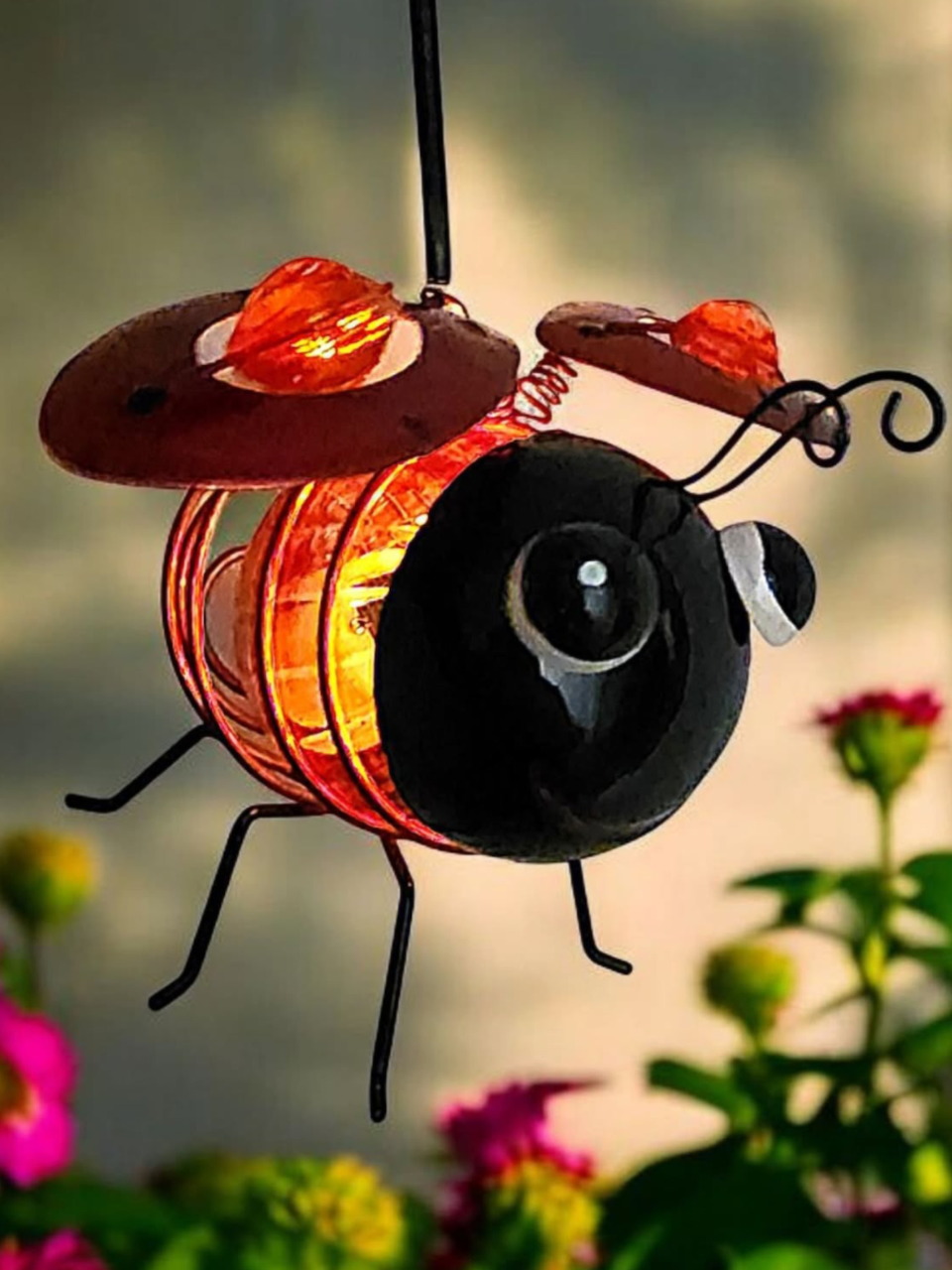 Bee-shaped hanging garden ornament with orange wings and flowers behind it