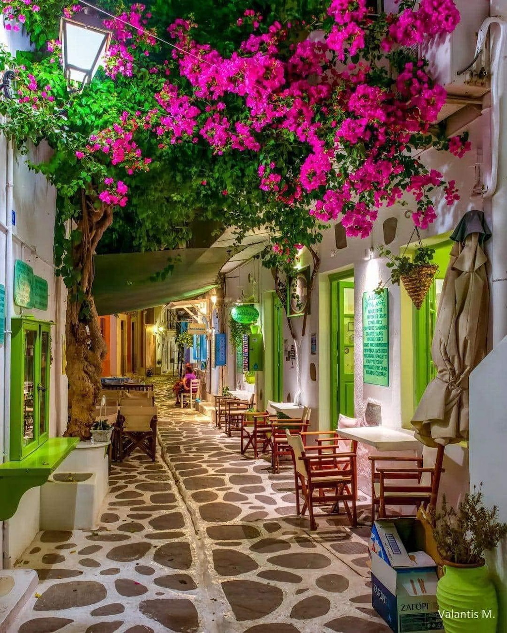 Cobblestone street in Paros with pink flowers, white buildings, green shutters, and cafe tables
