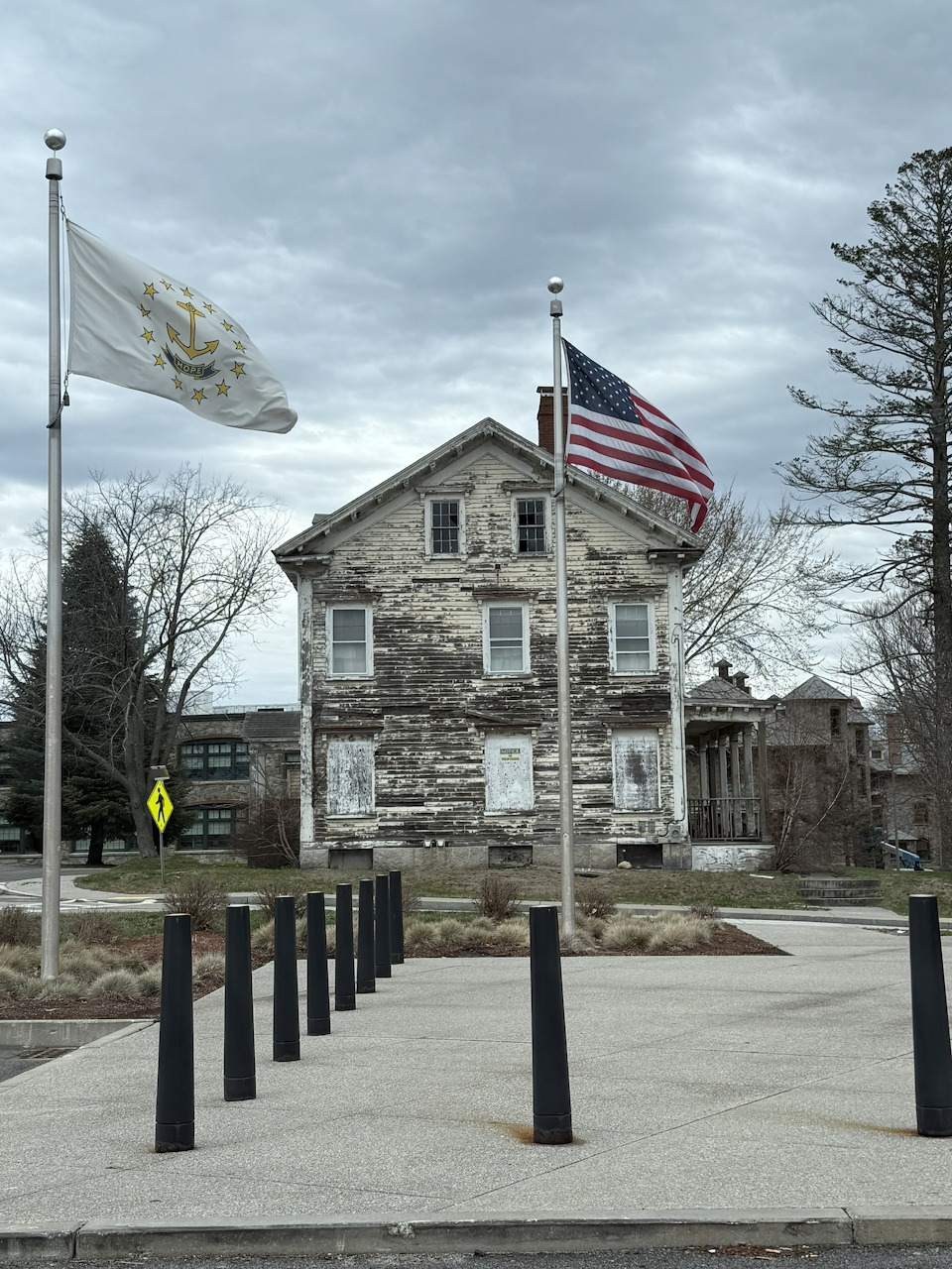 Old weathered stone building with American and state flags on poles, bollards in foreground, overcast sky