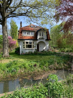 Small white cottage with red tiled roof beside a pond surrounded by green trees and gardens
