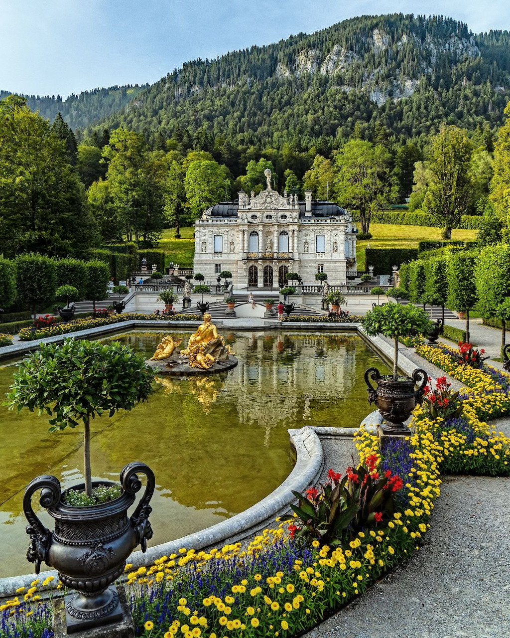 White baroque palace with ornate gardens, reflecting pools, golden fountains and forested mountain behind
