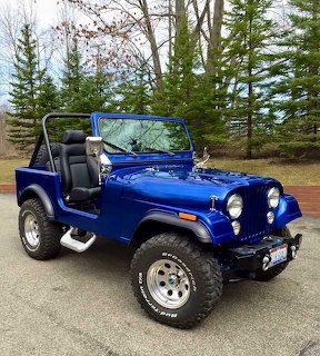 Blue open-top off-road Jeep parked on a driveway with trees behind it.