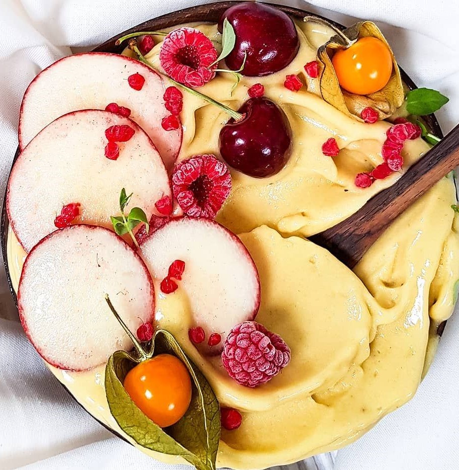 Yellow smoothie bowl with sliced apples, raspberries, cherries, and physalis on marble surface