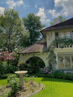 Two-story house with arched porch, climbing vines, birdbath in a green garden under blue sky