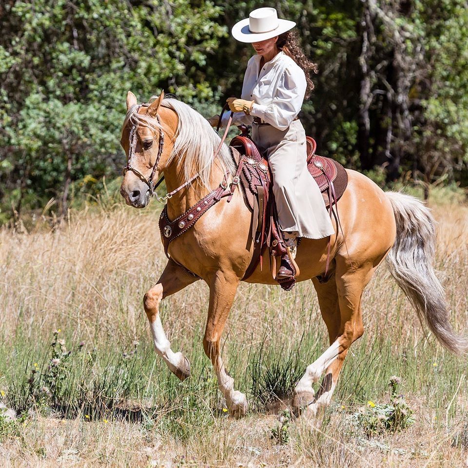 Woman in white hat and outfit riding a palomino horse through a grassy field