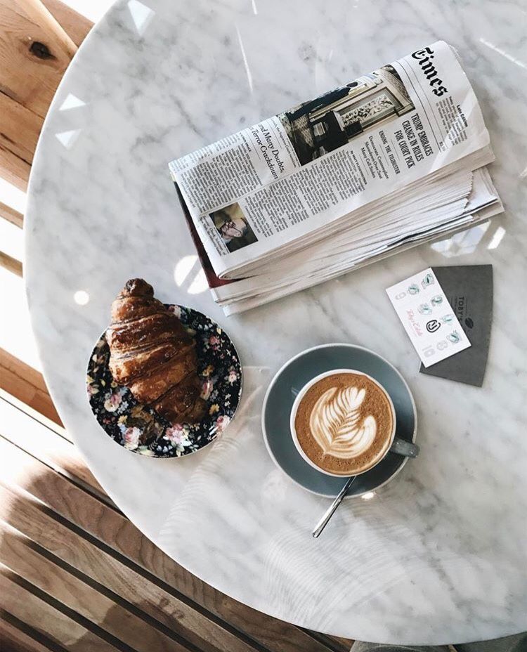 Latte with heart art, croissant on patterned plate, and newspaper on white marble table