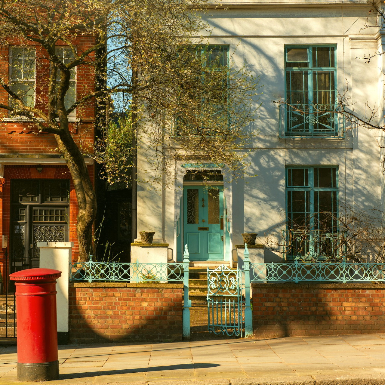 Sunlit townhouse facade with turquoise door, iron railings, trees, and a red postbox