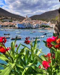 Mediterranean coastal village with colorful buildings, harbor boats, and pink flowers in foreground under cloudy sky