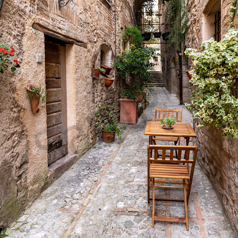 Narrow cobblestone alley in Spello, Italy, with wooden chair, potted plants, and stone buildings